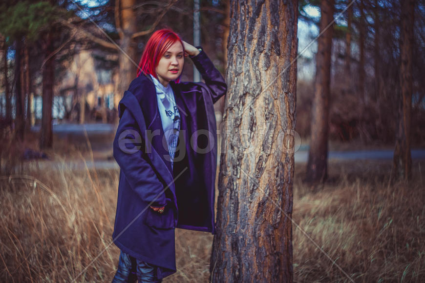 Девушка с ярко-красными волосами стоит, опираясь рукой на дерево