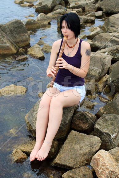 Девушка с татуировкой играет на японской медитативной флейте сякухати, сидя на камнях